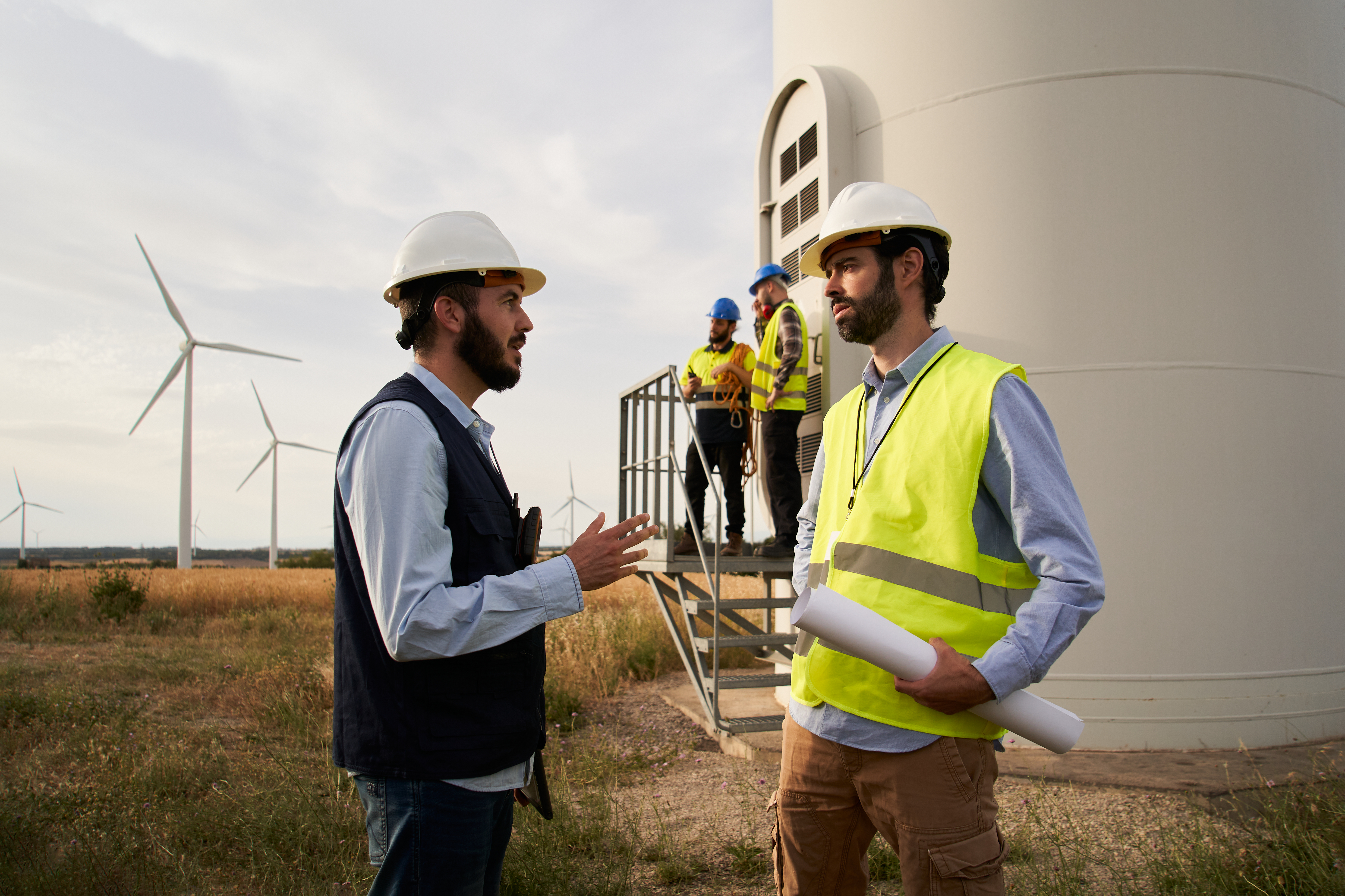 Group Of Engineers In Wind Power Station. Renewable And Clean Energy Concept, Sustainable Future. Group of engineers in hard hats talking and working with a blueprint in an agricultural field with wind turbines. Wind power station. Renewable and clean energy concept, sustainable future.